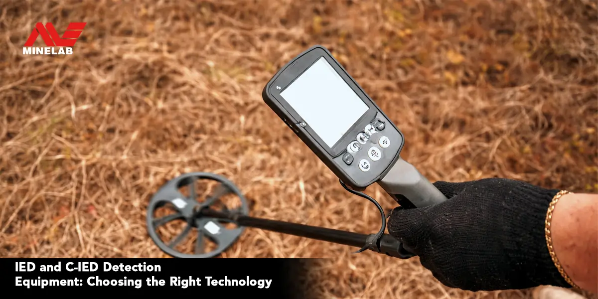 Handheld Minelab metal detector with digital display and search coil being used outdoors to detect buried metal objects in dry ground, illustrating IED and C-IED detection equipment technology.
